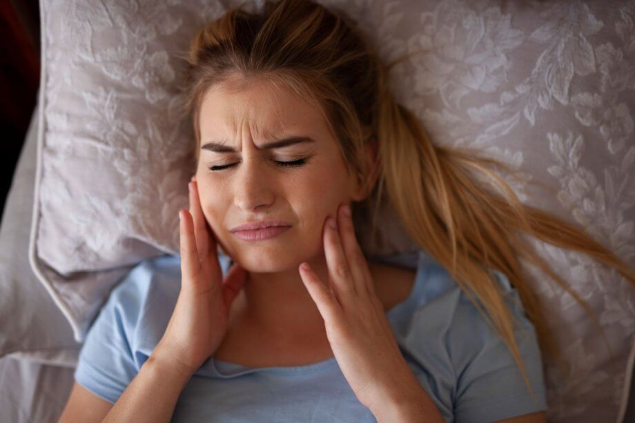 Blonde woman laying in bed on white pillows holding jaw in pain